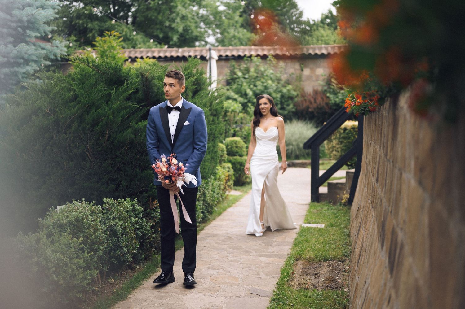 A groom in a navy suit holds a bouquet while a bride in a white gown walks toward him in a lush garden.