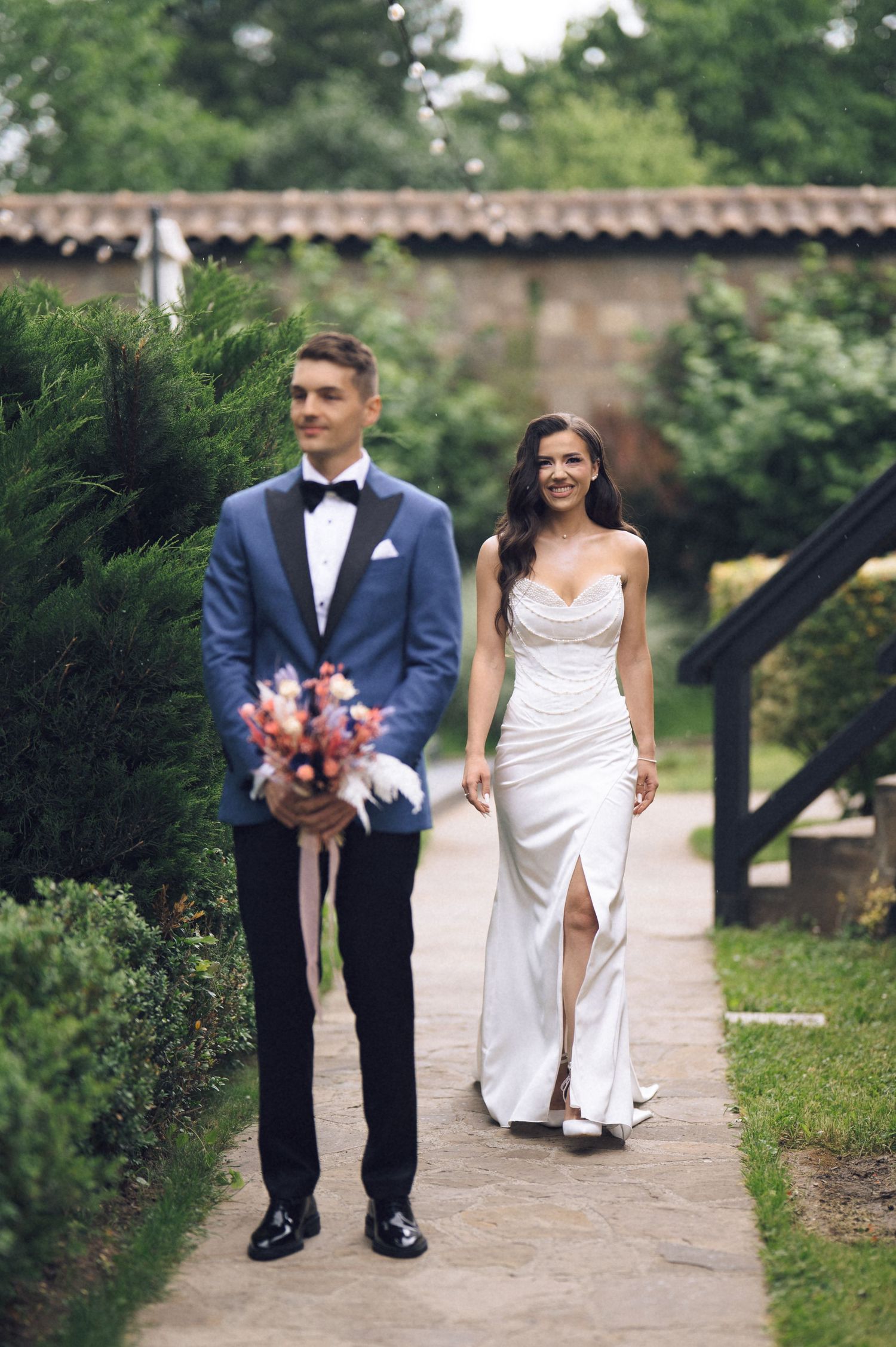 Bride in white gown and groom in blue tuxedo walking together on a garden pathway at their wedding.