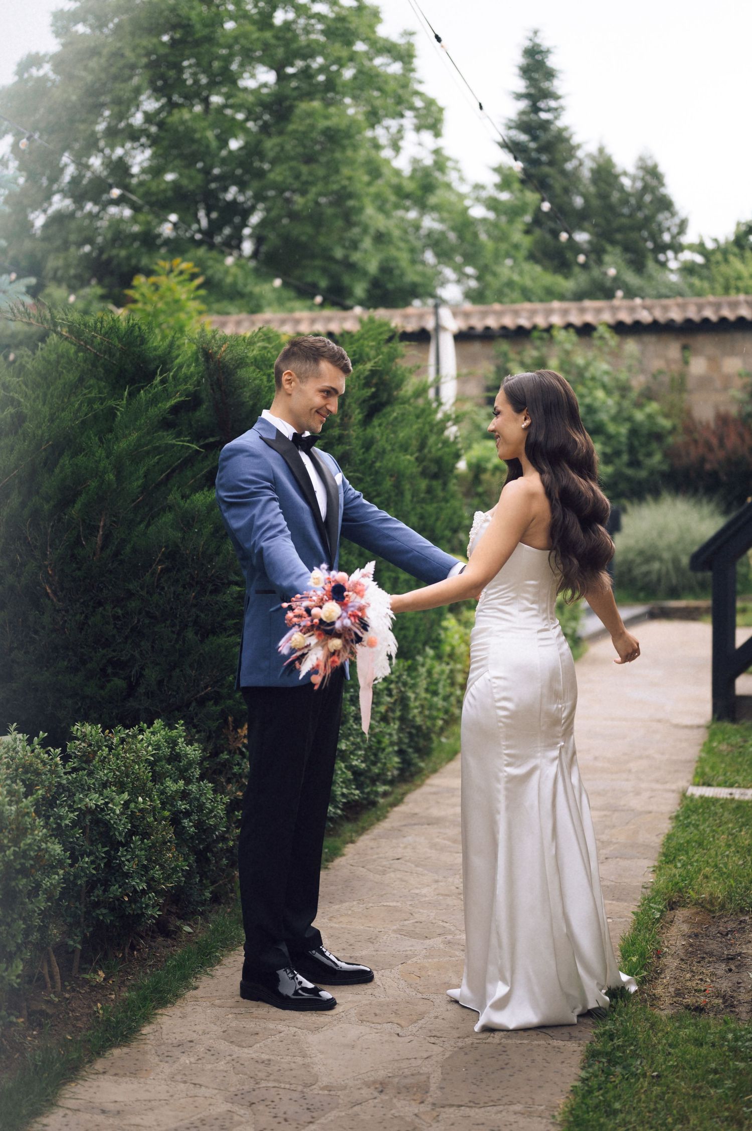 Bride in white gown and groom in blue tuxedo hold hands in a lush garden setting on their wedding day.