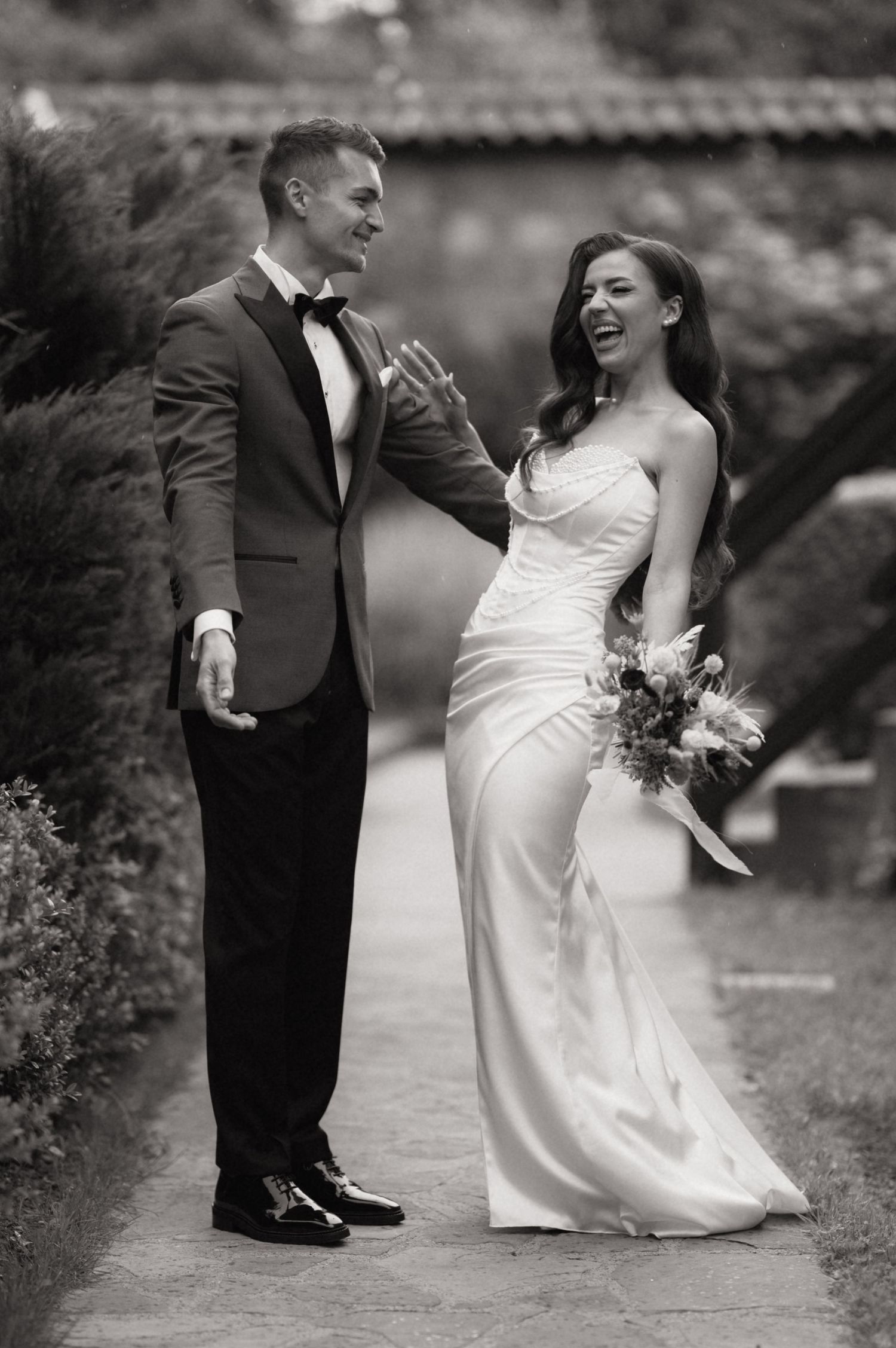 Black and white photo of a laughing bride in a sleek gown and groom in a tuxedo sharing a joyful moment outdoors.