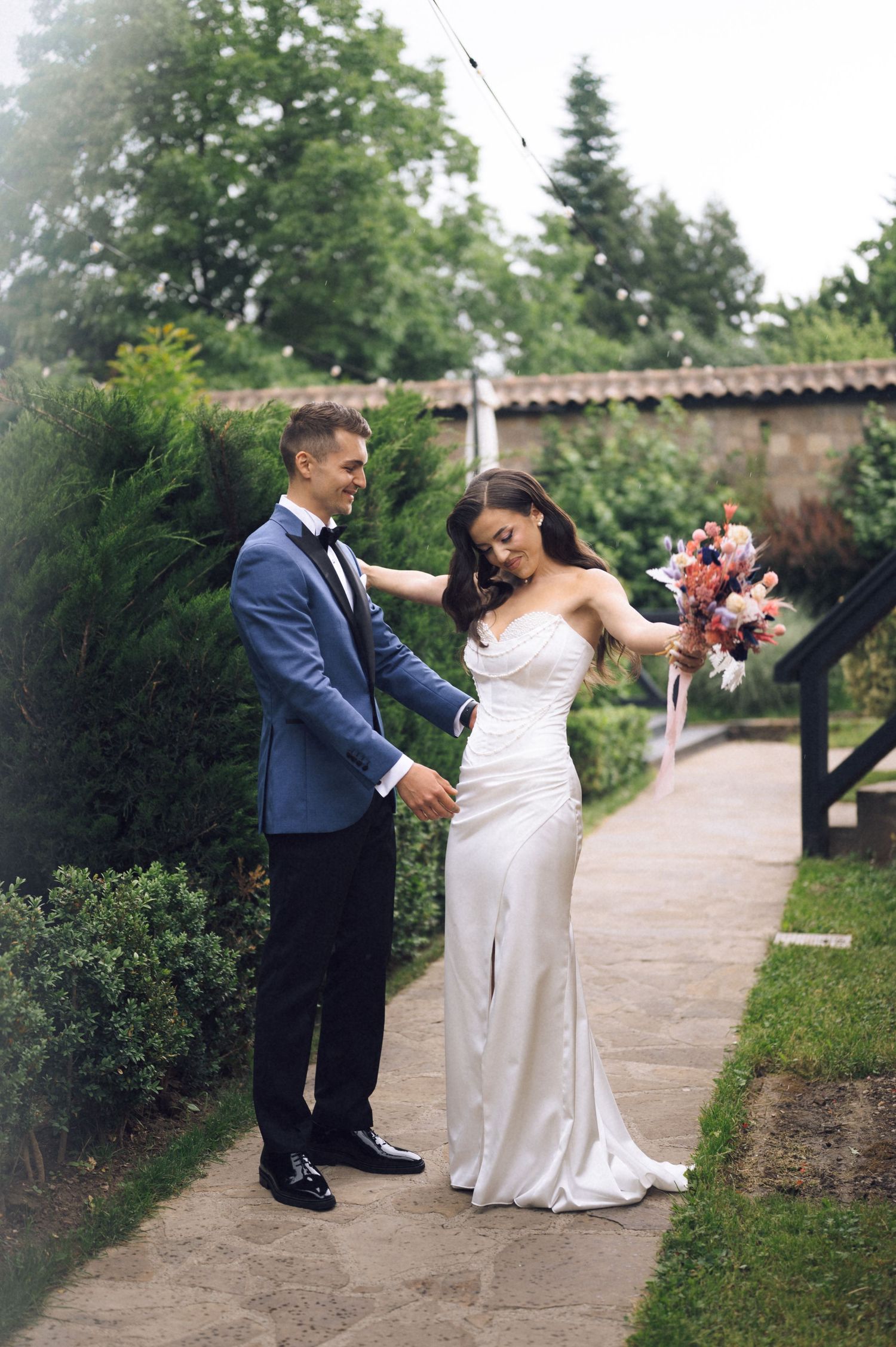 Bride in white gown holds bouquet as groom in blue suit tenderly holds her hand on a garden pathway.