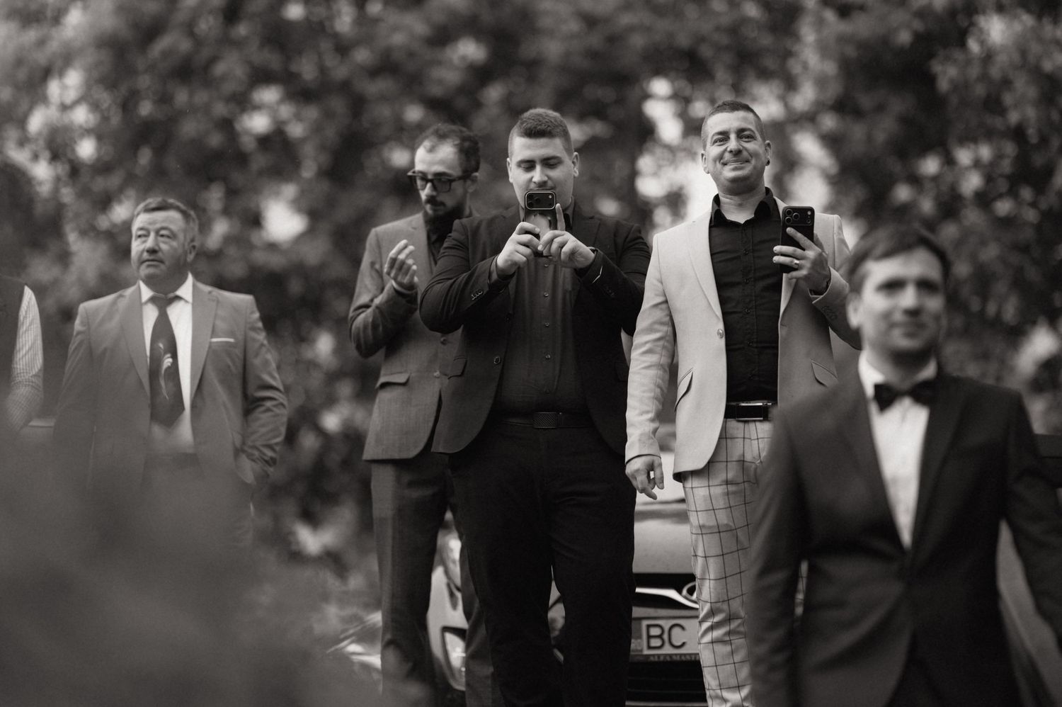 Black and white photo of four men in suits outdoors, two using smartphones near a parked car.