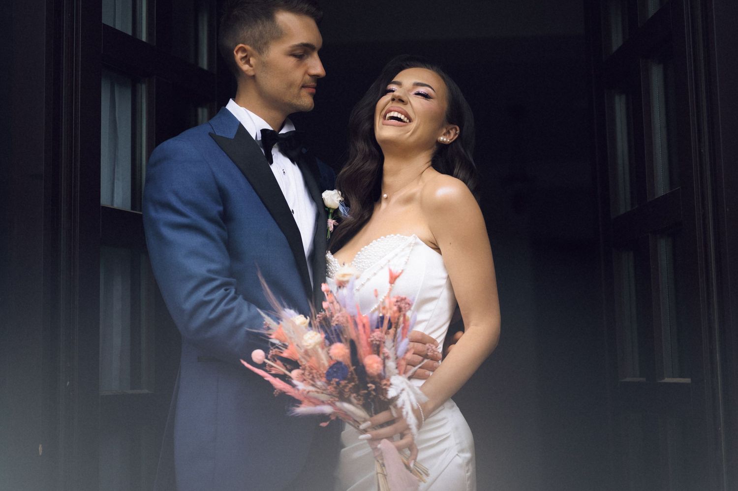 Bride and groom share a joyful moment in a doorway, she in a white gown holding a bouquet, he in a blue tuxedo.