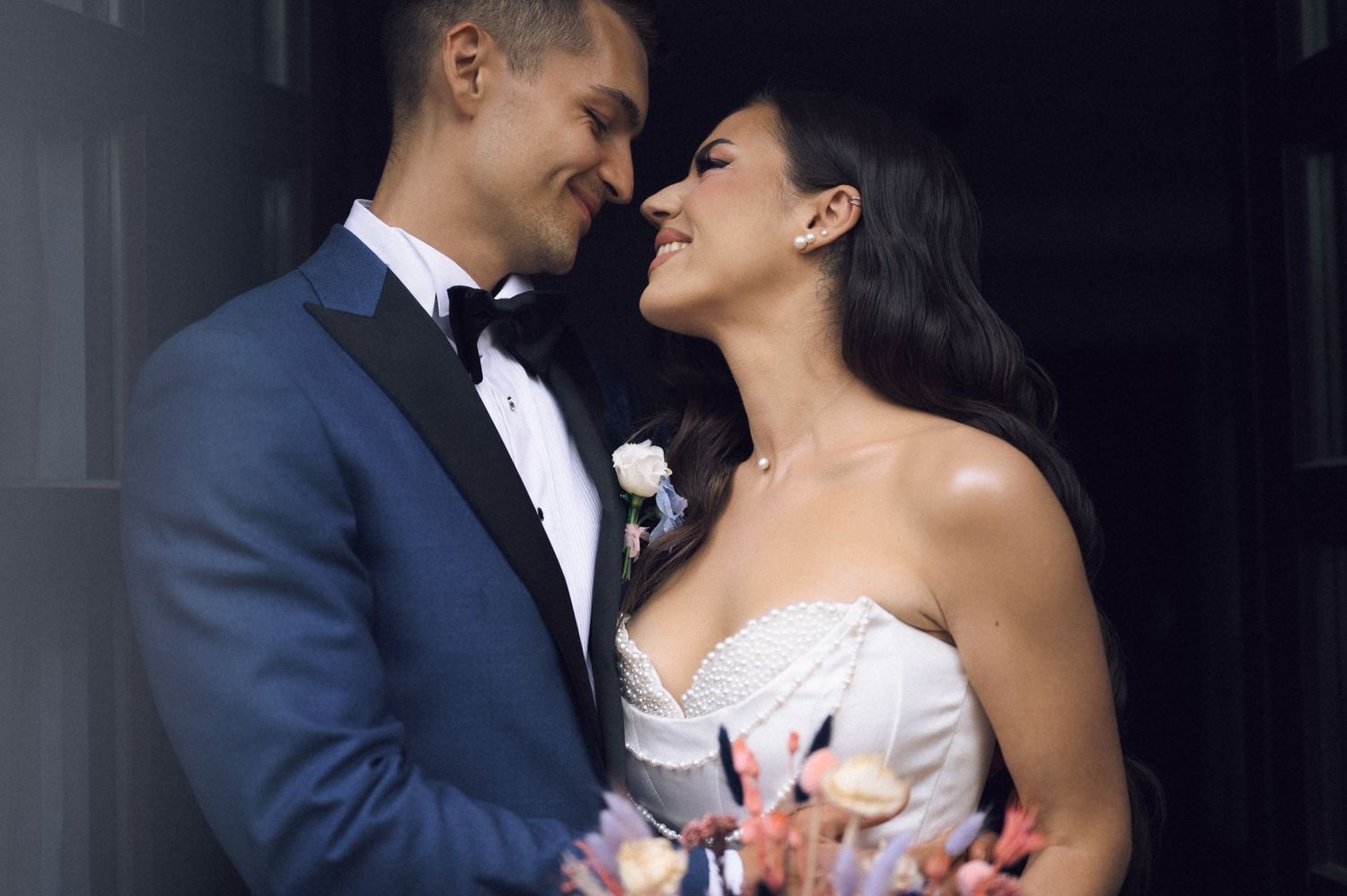 A bride and groom share a romantic moment, smiling at each other, she holds a bouquet, he wears a blue tuxedo.