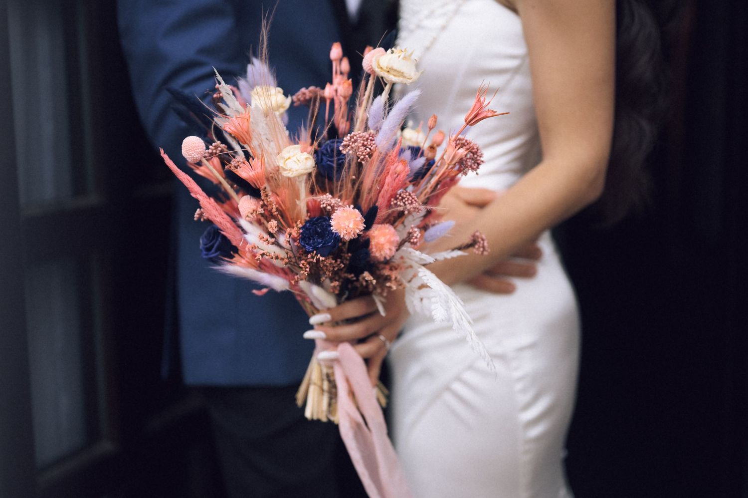 Bride in white dress holding a colorful dried flower bouquet with pink and purple blooms beside groom in blue suit.
