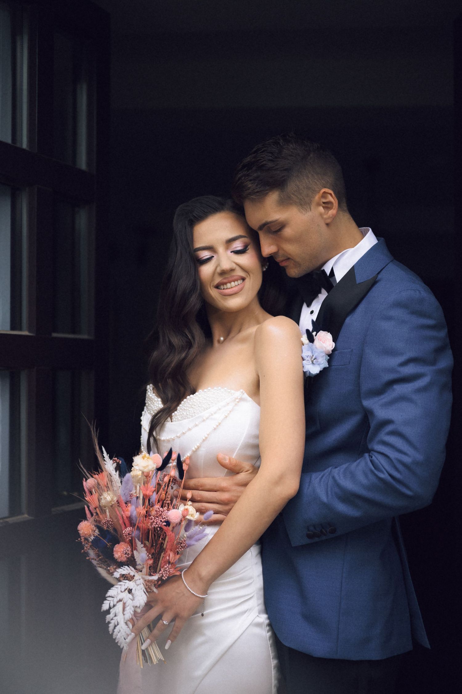 Bride in white gown holds colorful bouquet while groom in blue tuxedo embraces her tenderly near a dark window.