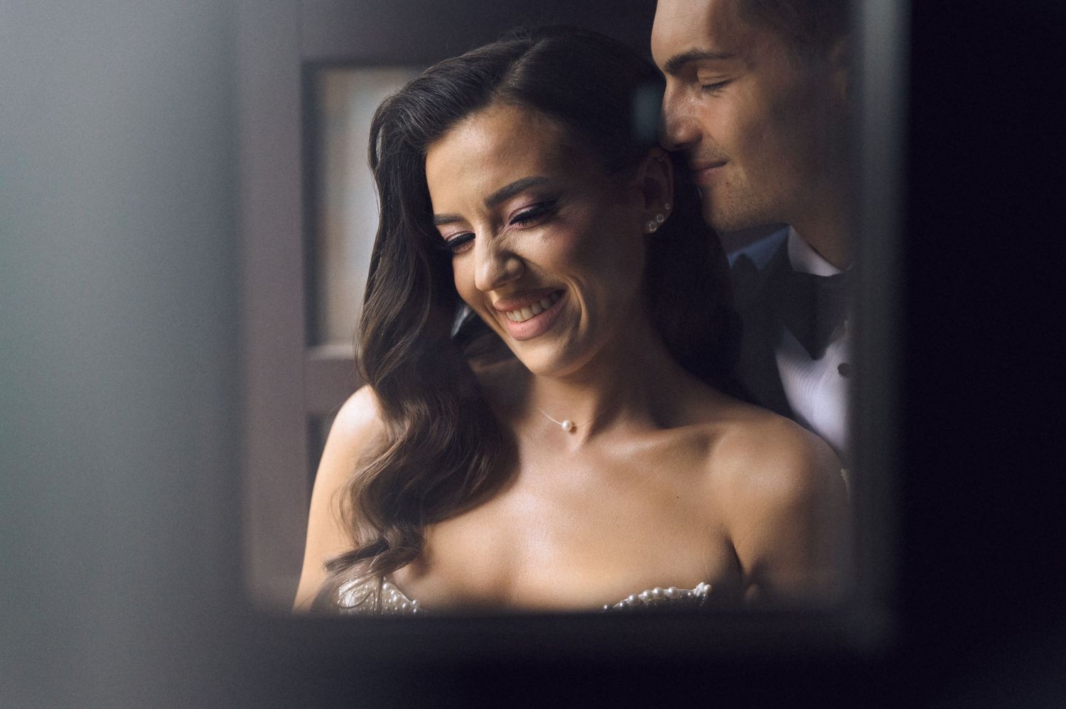 Elegant woman in strapless gown smiling softly at a formal event, with a man in a tuxedo behind her.