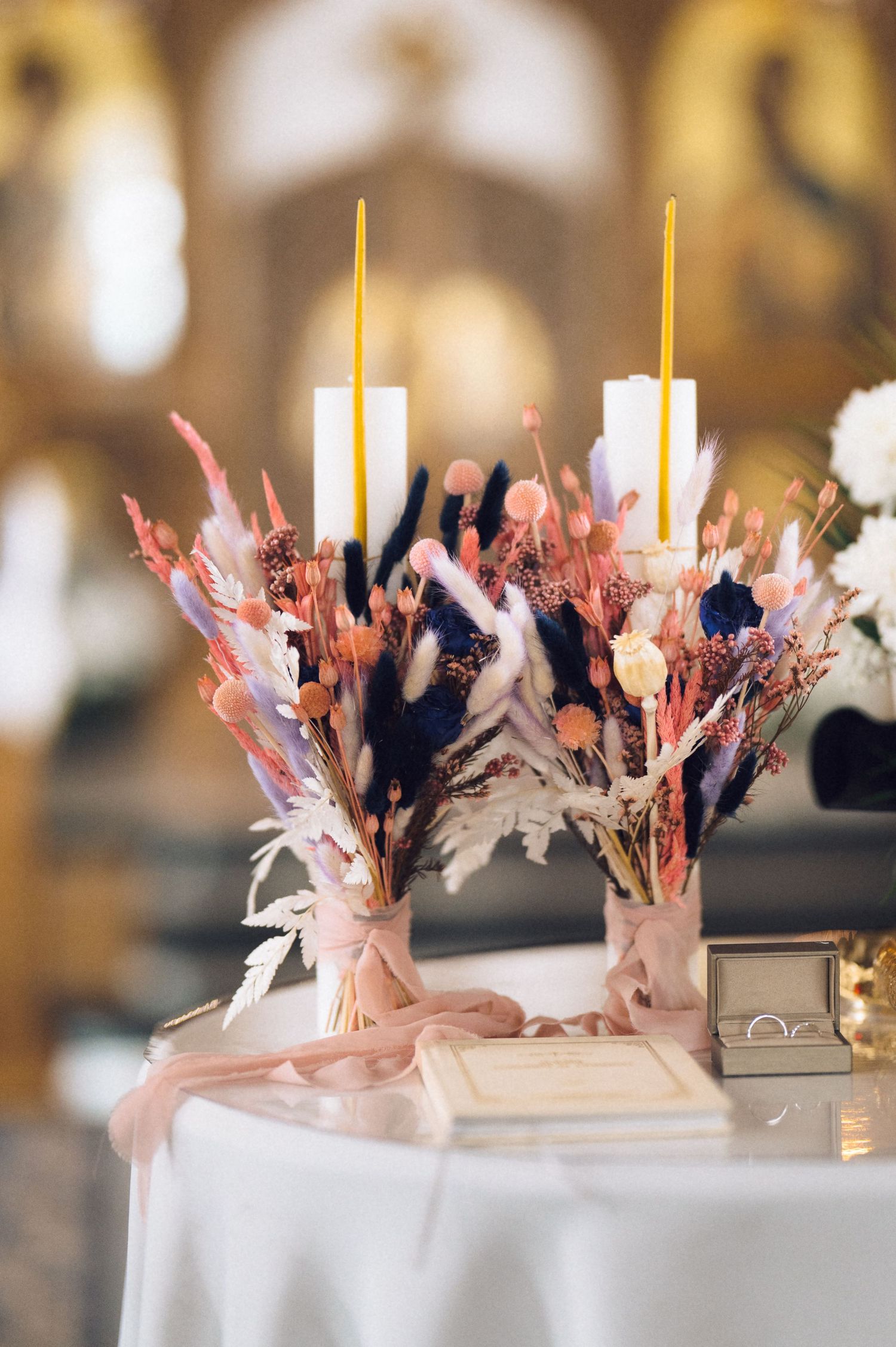 Elegant wedding table with dried floral arrangements, white and yellow candles, pink ribbon, and ring box in ornate venue.