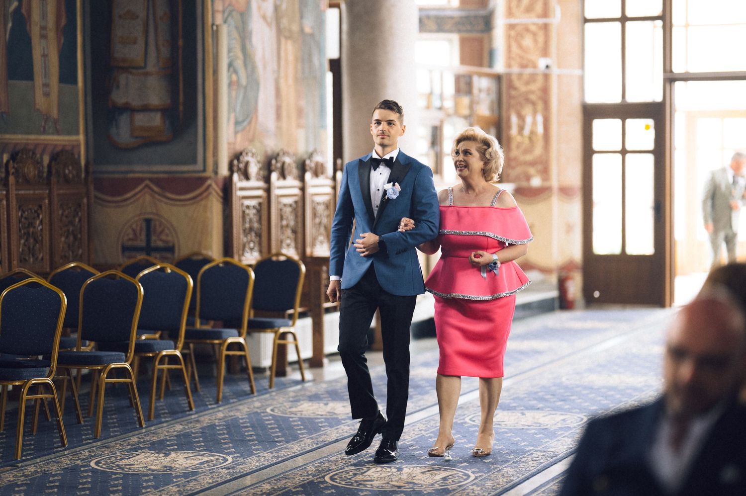 Groom in navy tuxedo walks with elegantly dressed woman in pink dress through ornate wedding venue aisle.