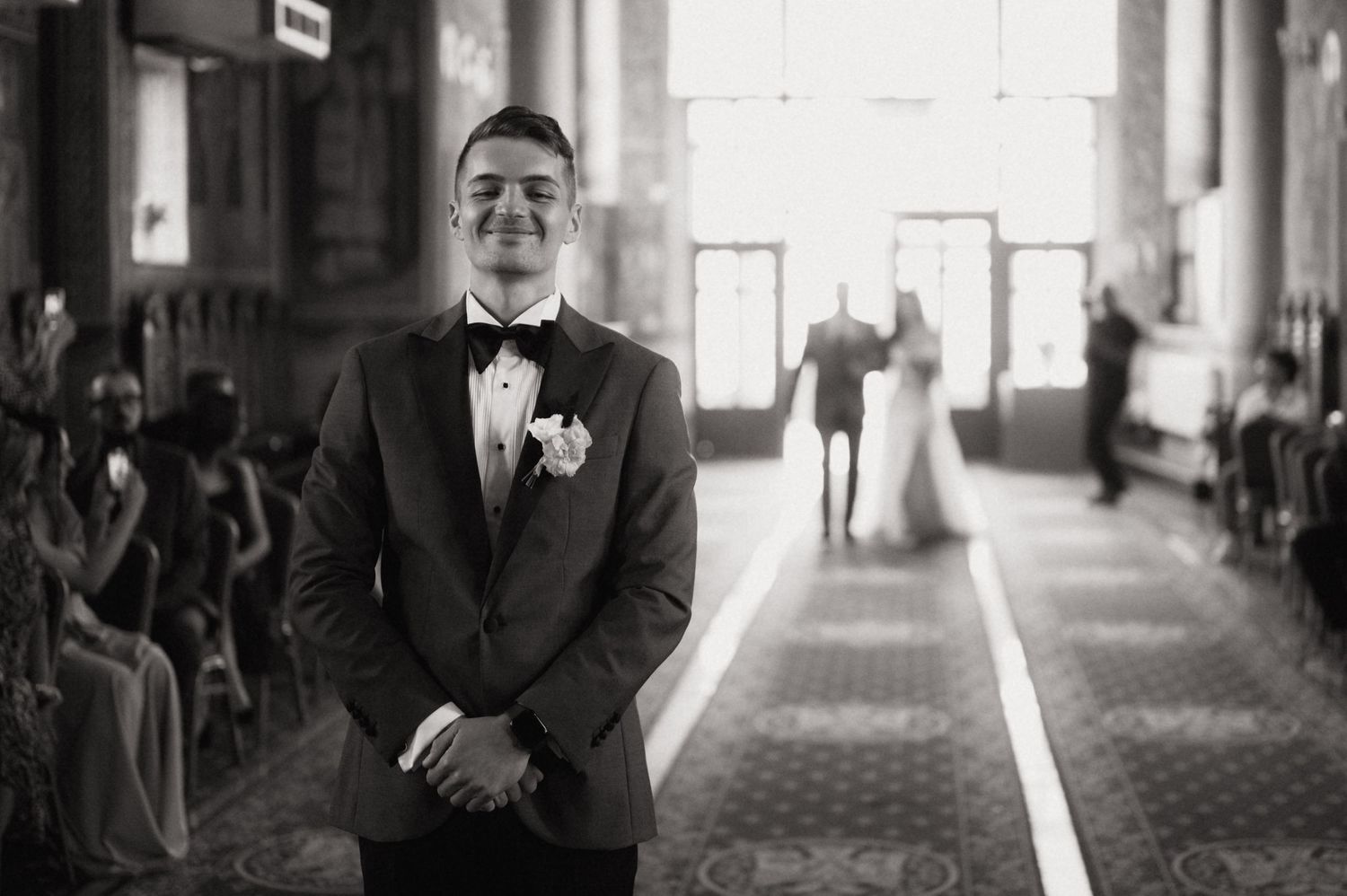 Groom smiling at altar in black tuxedo as bride walks down aisle in elegant black and white wedding photo.
