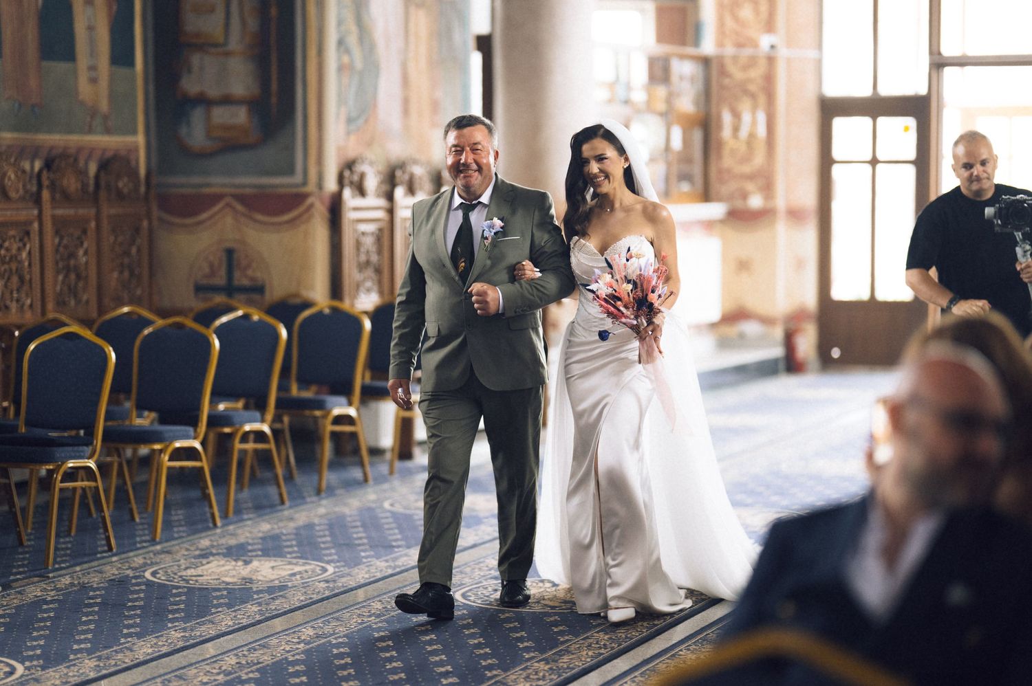 Bride walks down the aisle with her father in an ornate church during a wedding ceremony.
