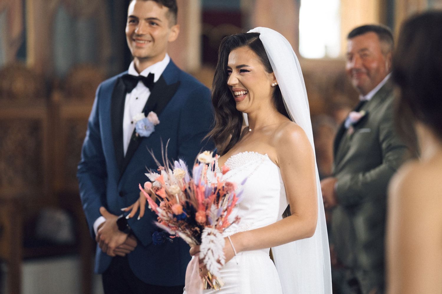 Smiling bride in white gown and veil holds pink bouquet at wedding ceremony with groom in blue tuxedo.