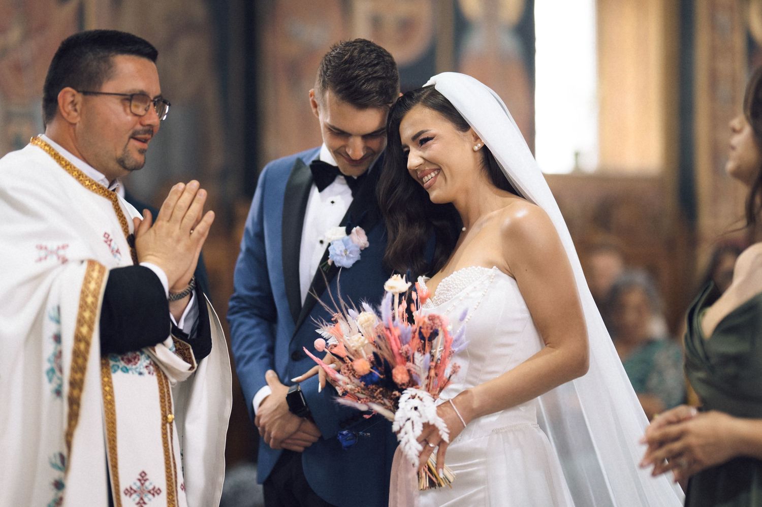 A joyful bride and groom share a moment with a smiling priest during a church wedding ceremony.