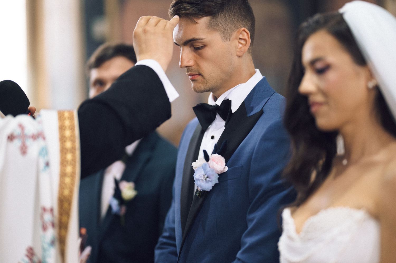 Groom in blue tuxedo making sign of cross during wedding ceremony at church altar with bride in white gown.