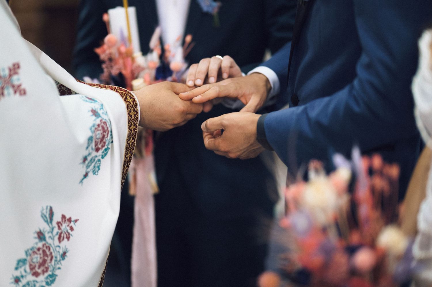 Officiant places wedding ring on brides hand during ceremony with floral decorations in background.