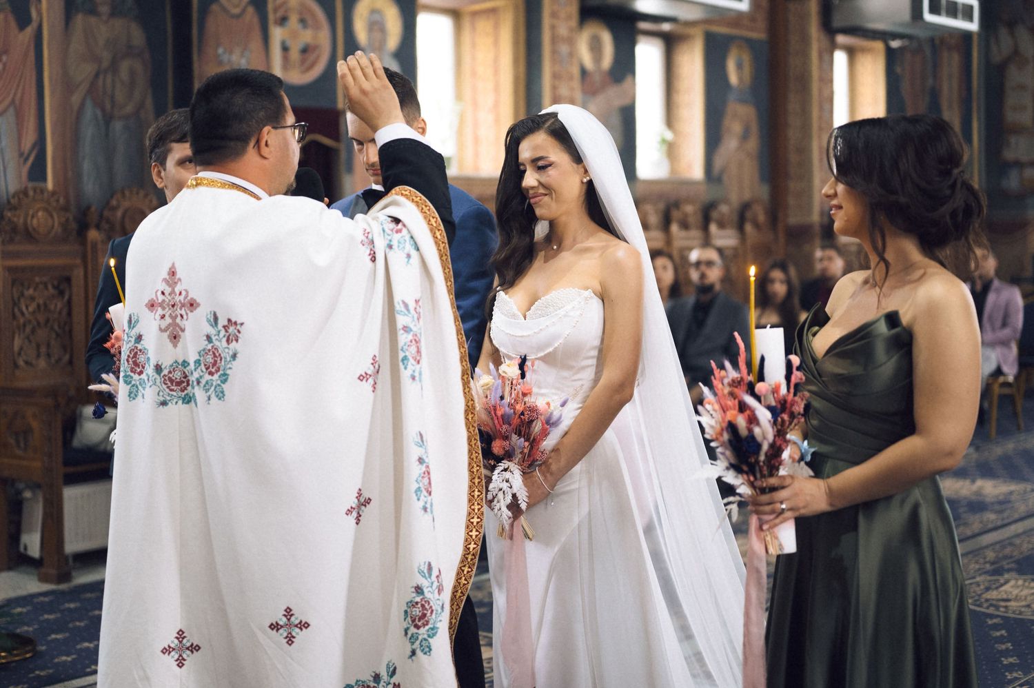 Bride in white gown receives blessing from Orthodox priest during church wedding ceremony with bridesmaid holding candle.