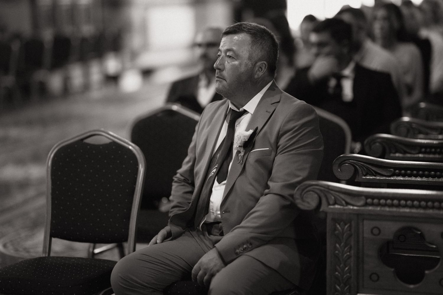 Black and white photo of a man in a suit sitting alone at a wedding ceremony, looking thoughtful.