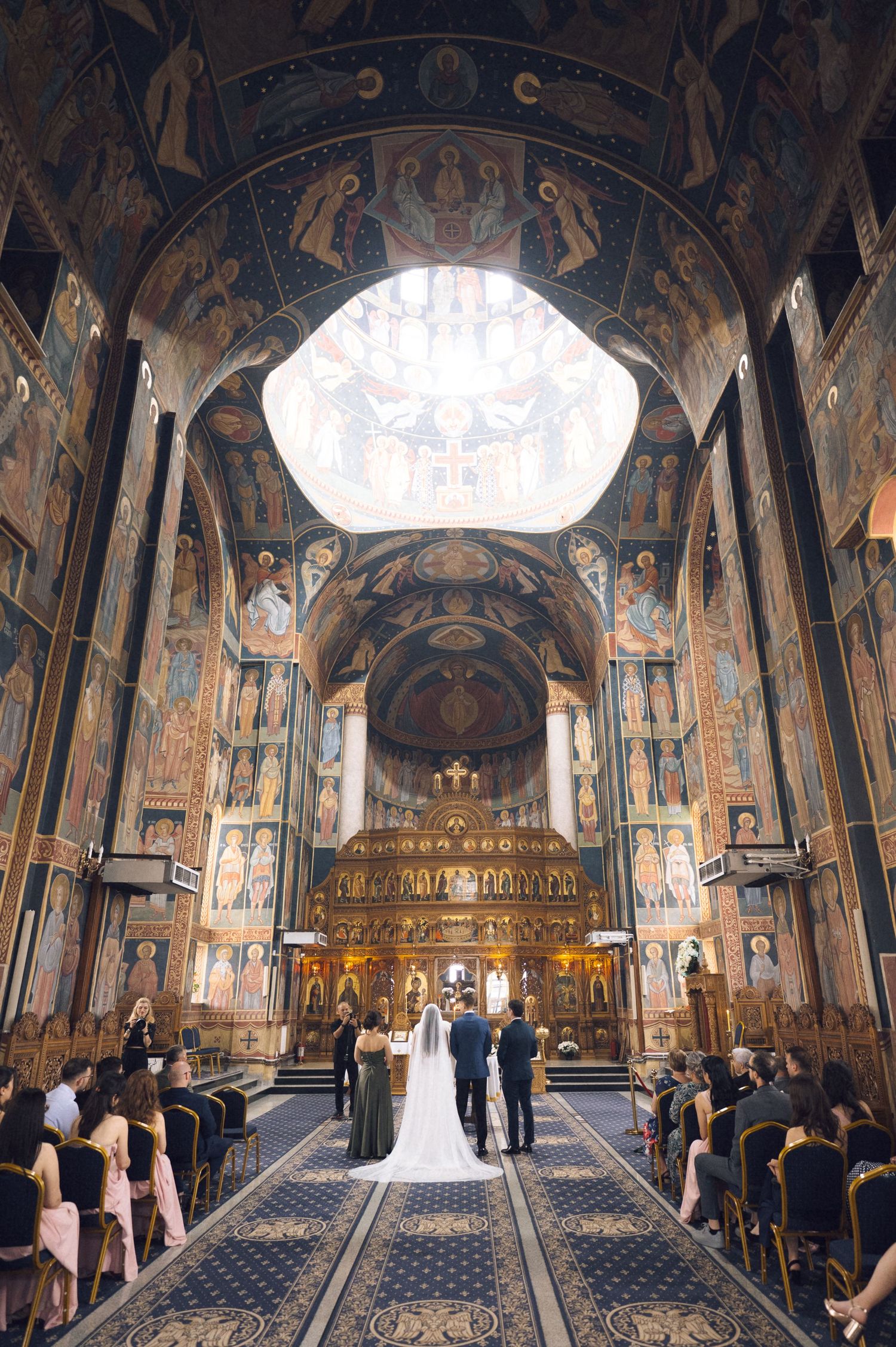 Elegant wedding ceremony in an ornate Orthodox church with Byzantine frescoes, grand arched ceilings, and a glowing central skylight.