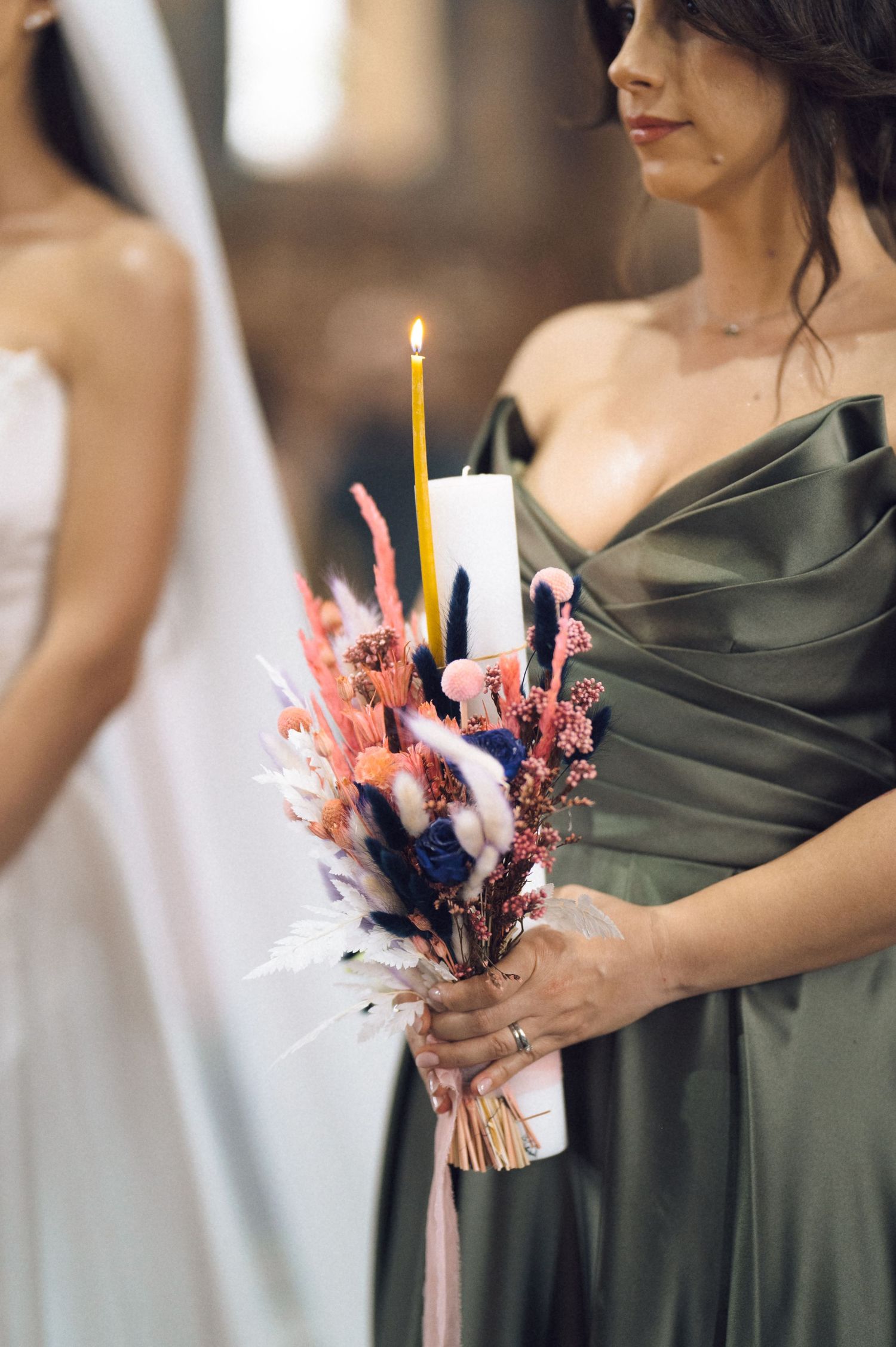 Bridesmaid in olive satin gown holding colorful dried flower bouquet with lit taper candle at wedding.