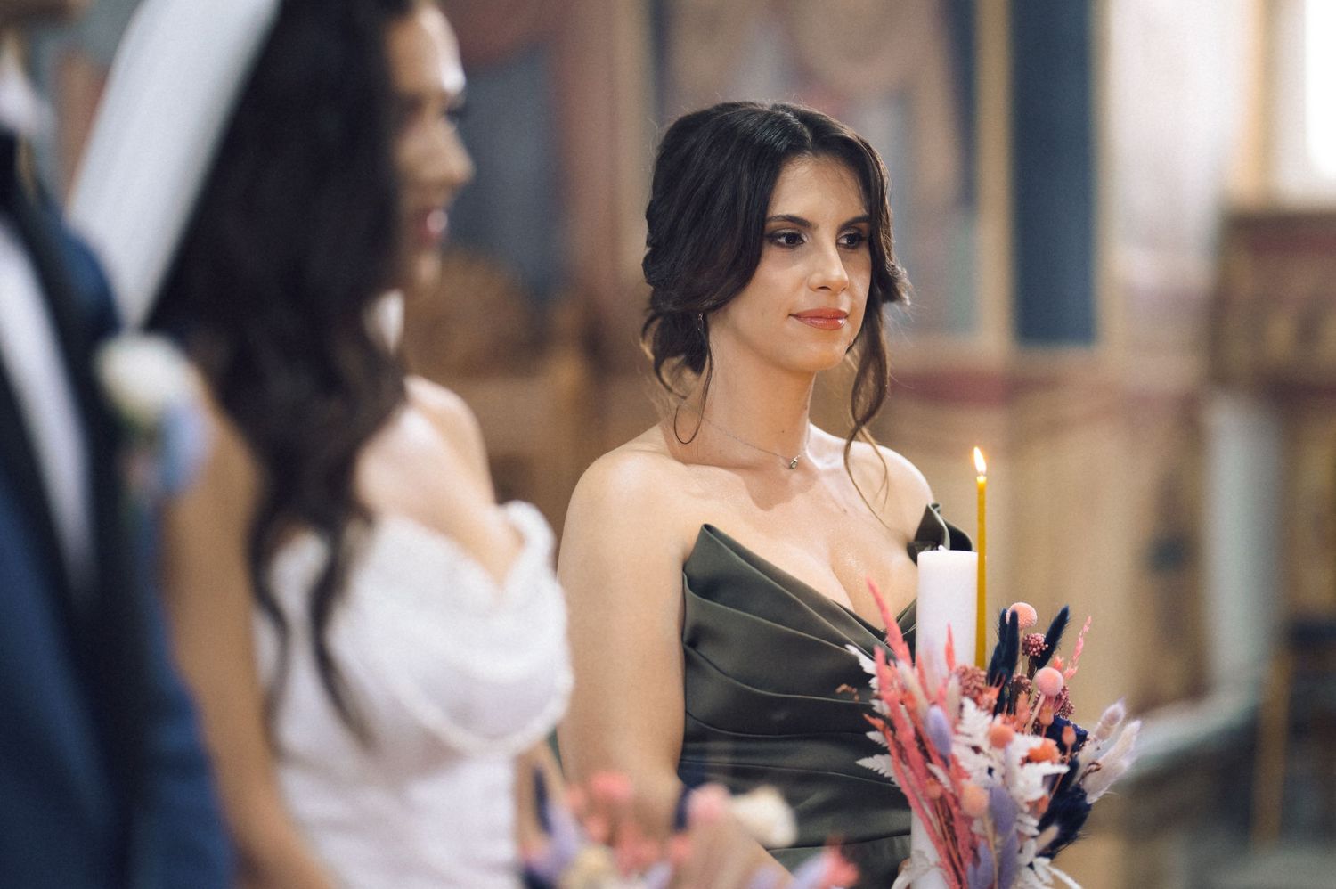 Bridesmaid in black dress holding candle and flowers at a church wedding ceremony.