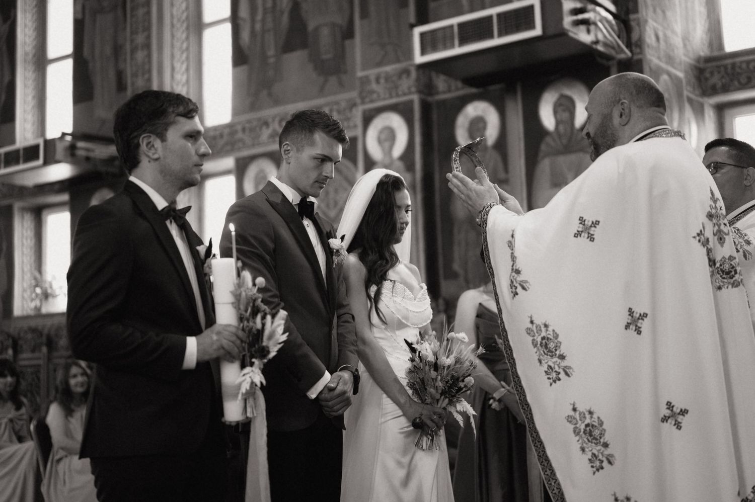 Black and white wedding ceremony photo of bride and groom with priest in ornate church.