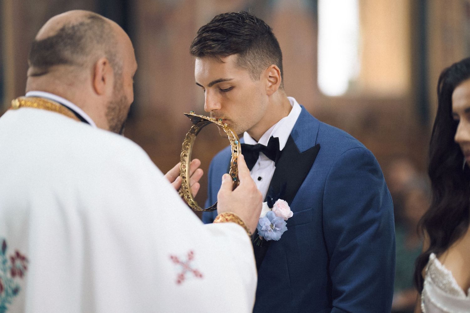 Groom in blue suit kissing ceremonial cross held by priest during traditional church wedding ceremony.