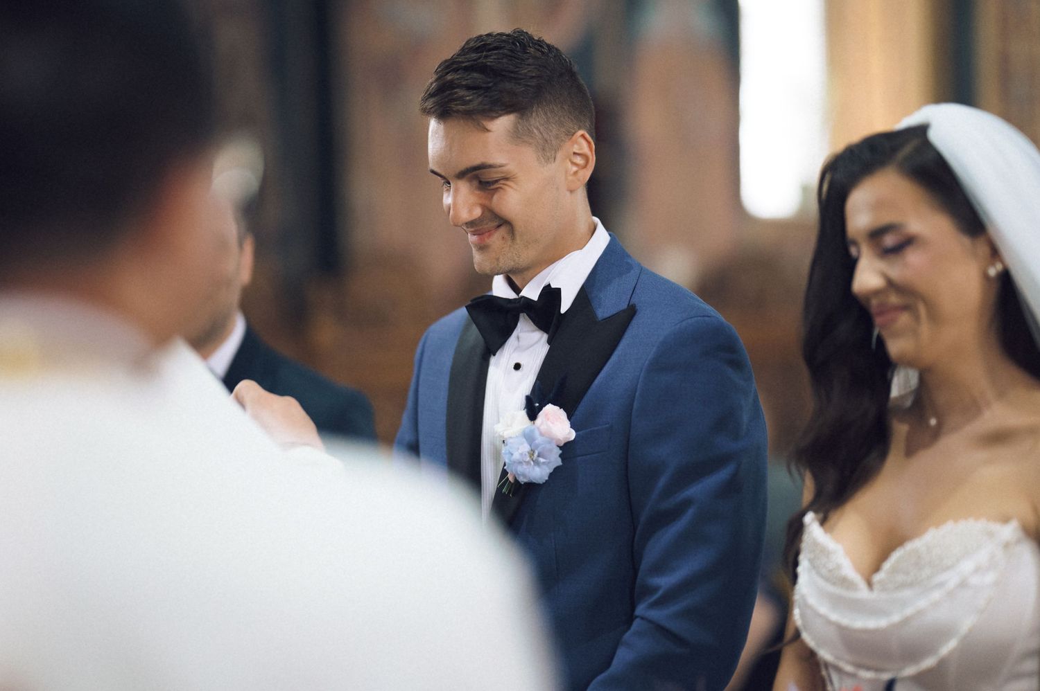 Smiling groom in blue tuxedo with bow tie stands beside bride in white wedding dress during church ceremony.