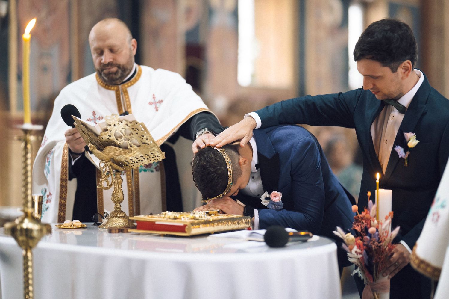 Groom bowing head during Orthodox wedding ceremony while priest reads scripture at ornate altar.