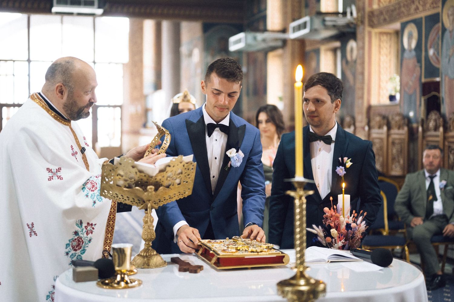 Two grooms in blue suits participate in a traditional Orthodox wedding ceremony with a priest at the altar.