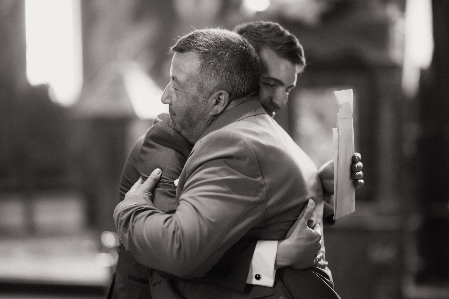 Two men in suits share an emotional hug at a formal event, one holding papers, in black and white.