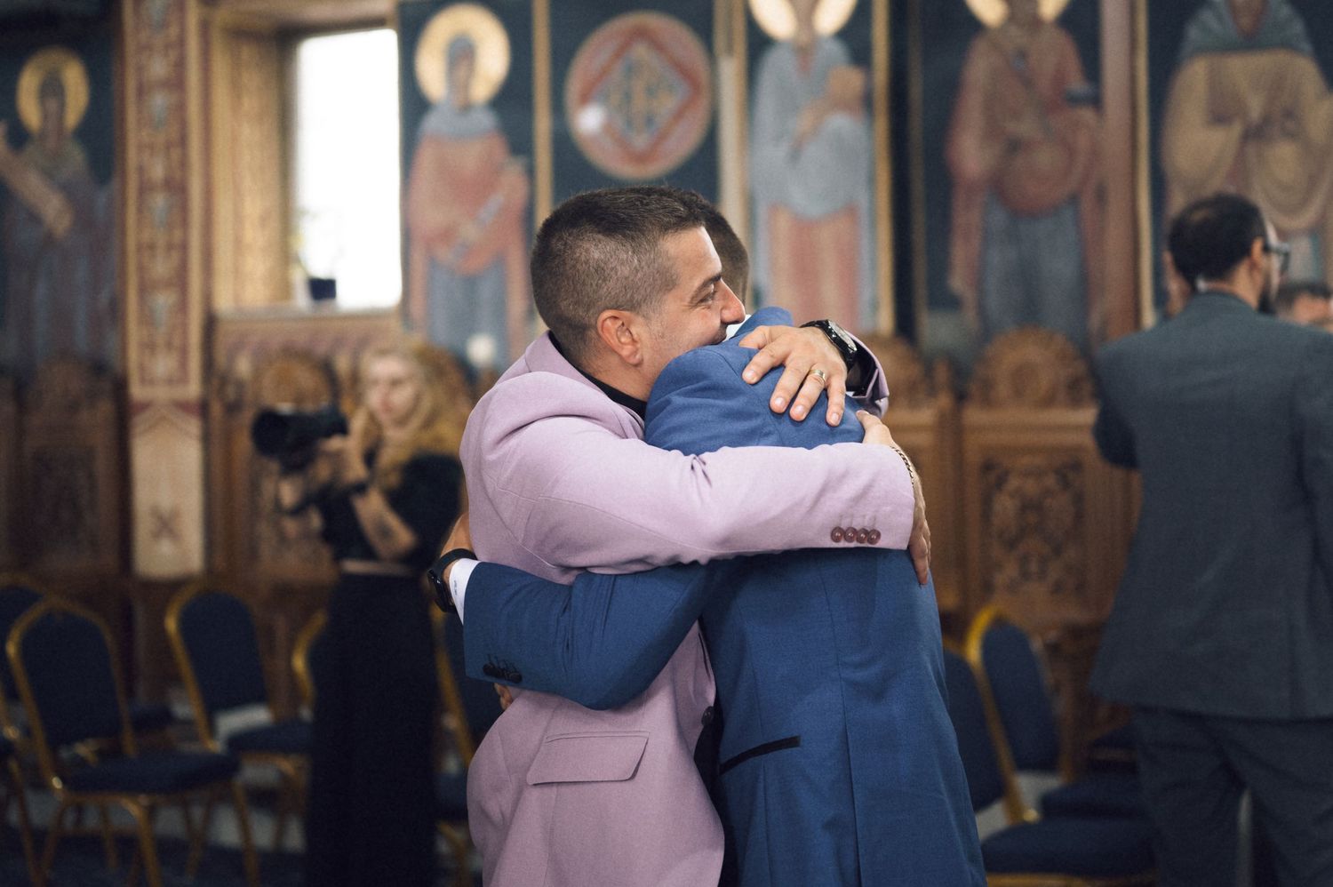 Two men in suits share a warm hug inside an ornate church during a formal ceremony.