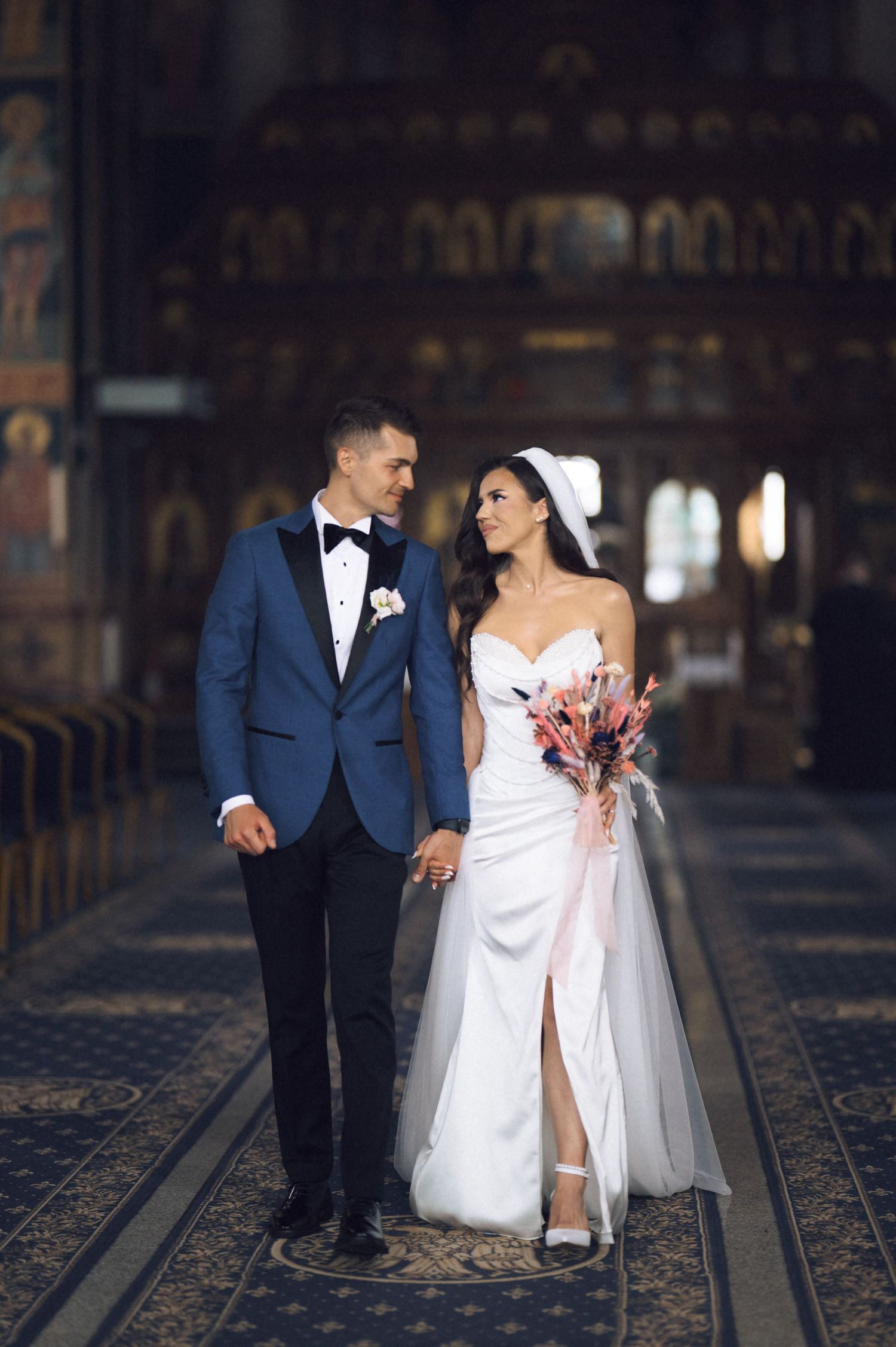 A bride and groom walk hand in hand inside an ornate church, she in a white gown, he in a blue tuxedo.