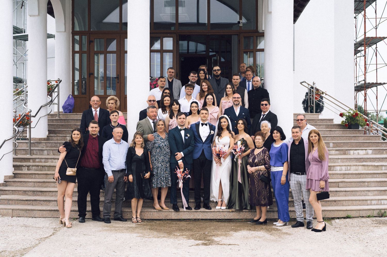 Large wedding party posing on grand building steps, bride and groom centered among formally dressed guests.