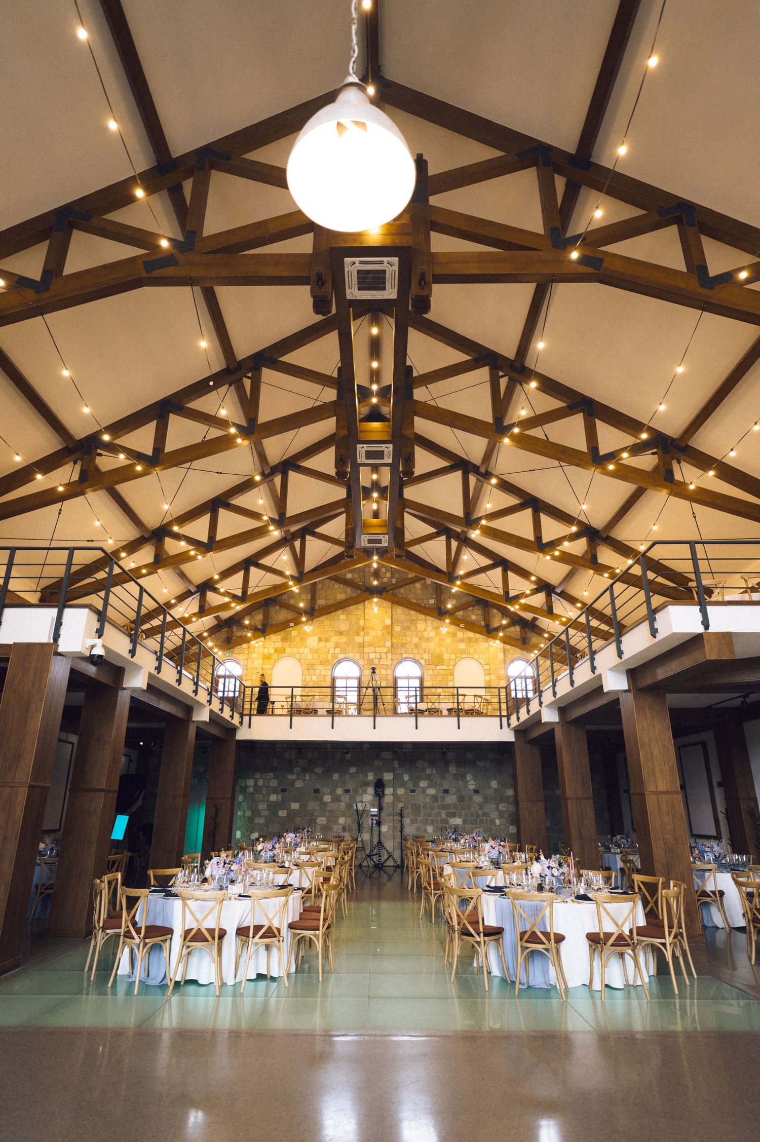 Elegant wedding reception hall with wooden beam ceiling, string lights, round tables with white linens, and warm ambient lighting.
