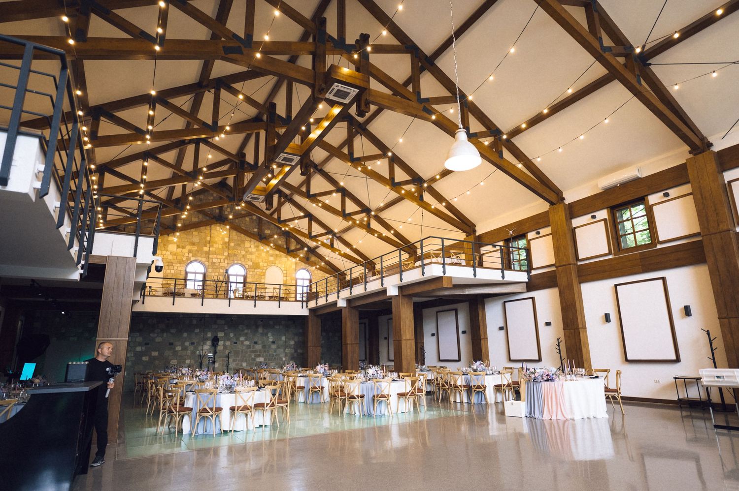 Elegant wedding reception hall with wooden beam ceilings, string lights, round tables, and a spacious dance floor.