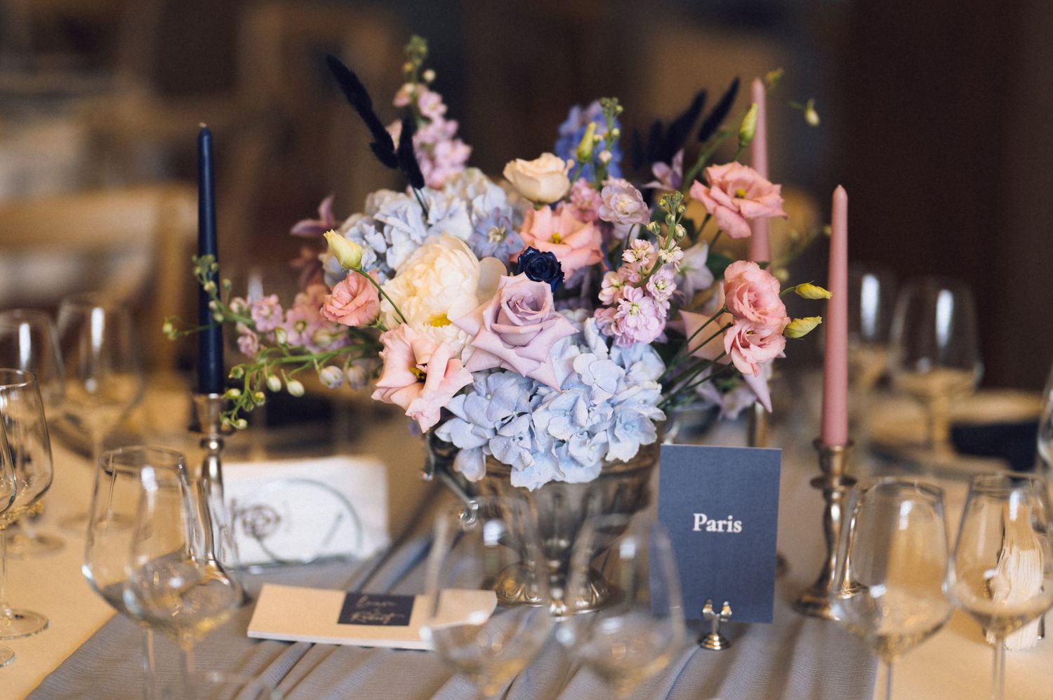 Elegant wedding table centerpiece with pink and blue hydrangea flowers, candles, and a Paris place card at a reception.