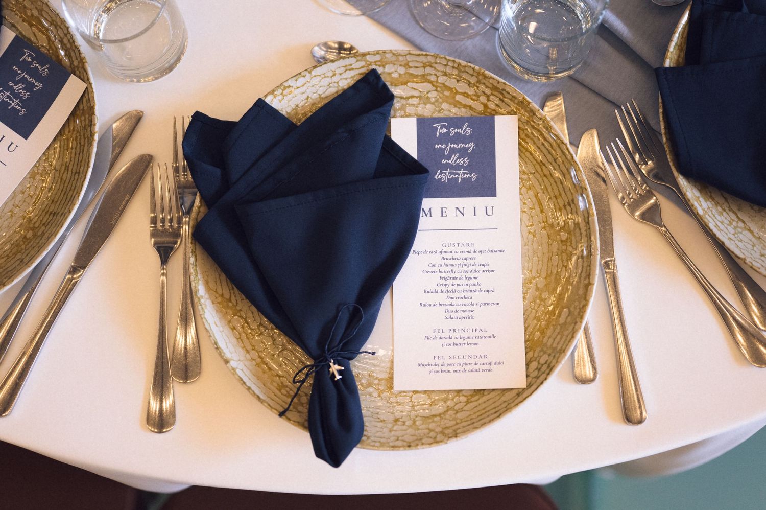 Elegant wedding place setting with navy blue napkin, gold charger plate, silverware, and a menu card on white tablecloth.