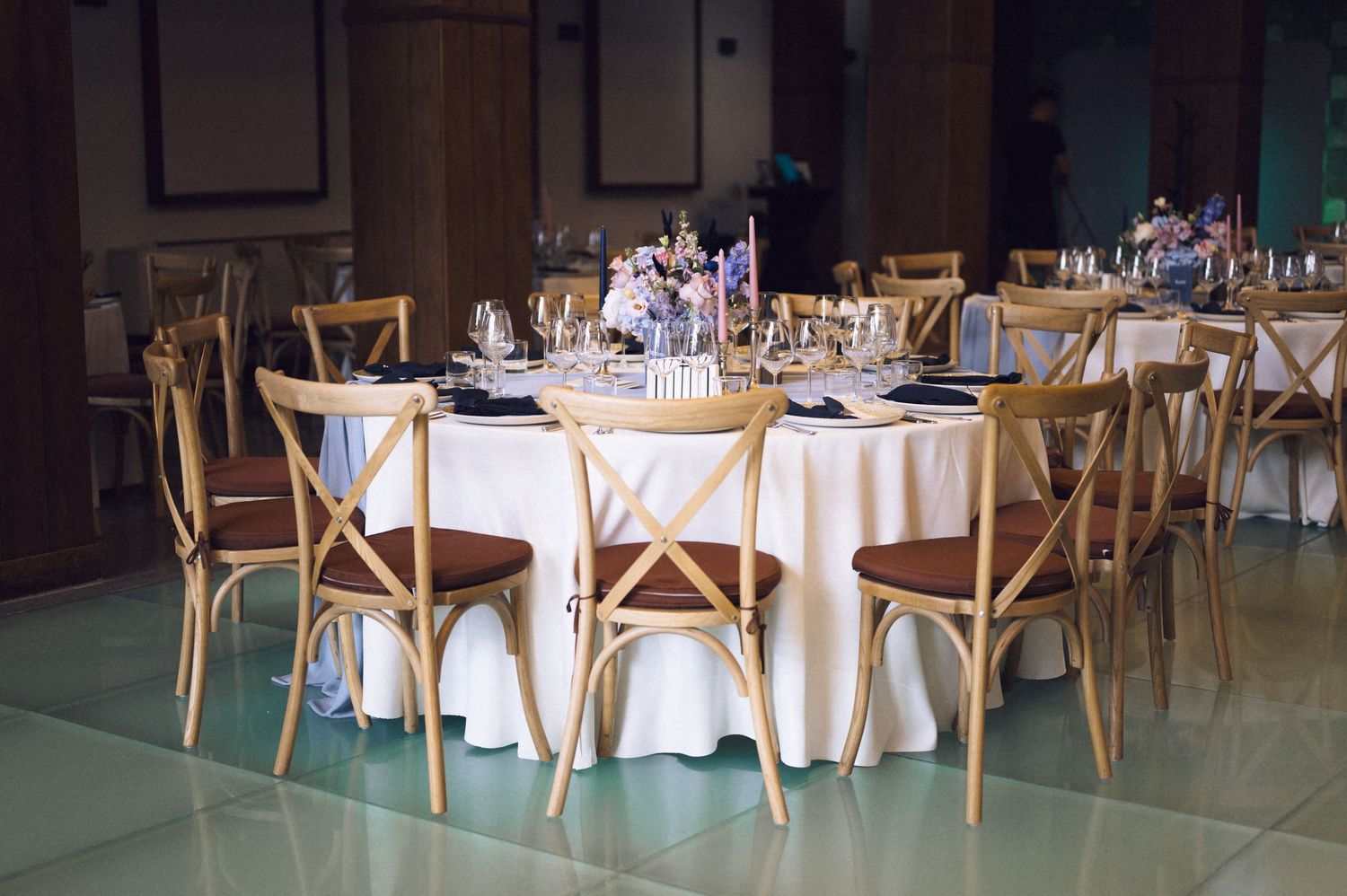 Wedding reception tables with white linens, wooden cross-back chairs, floral centerpieces, and elegant place settings in a hall.