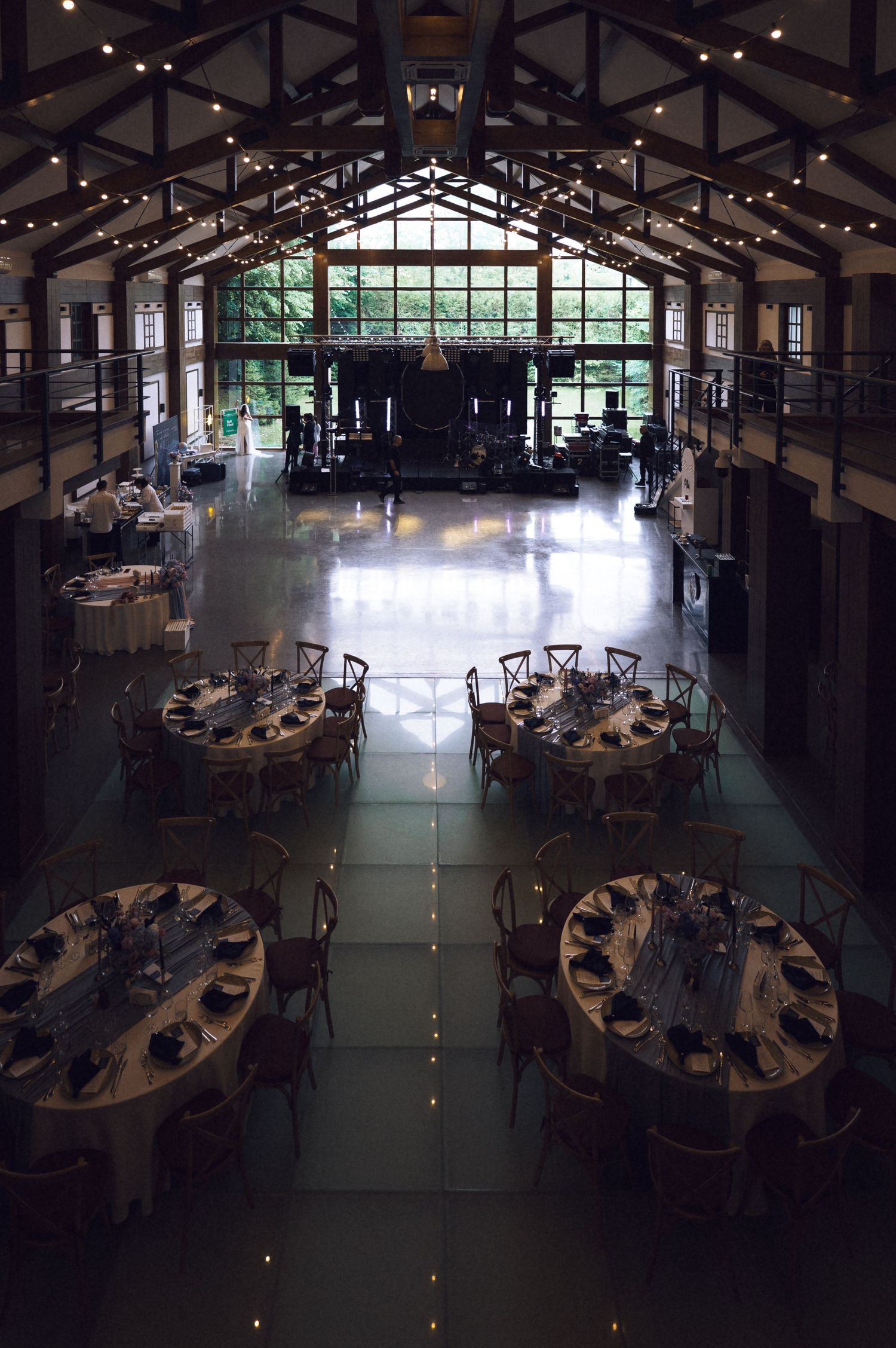 Elegant wedding reception venue with round tables, string lights, and a stage in a rustic barn-style hall.