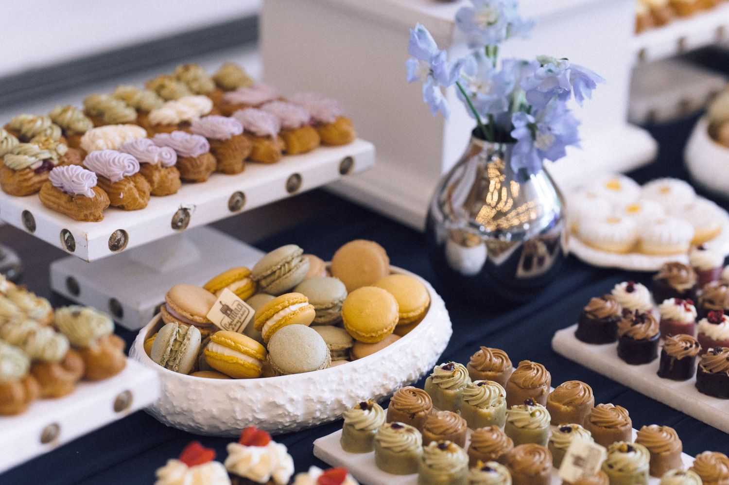 Elegant dessert table with colorful macarons, petit fours, and assorted pastries arranged on tiered white stands.
