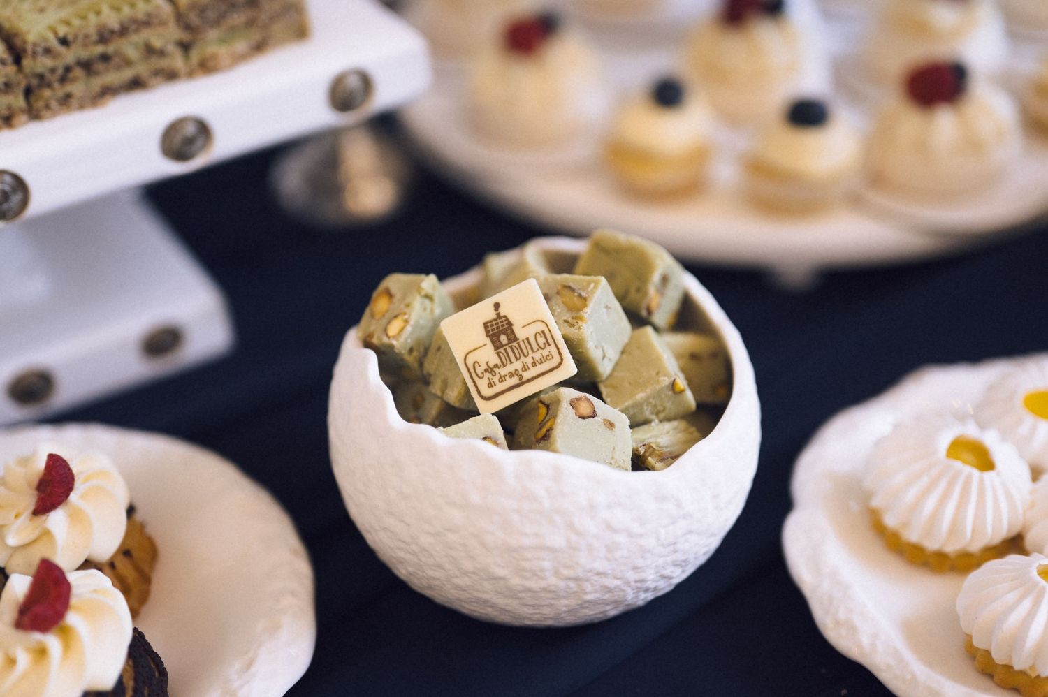 Elegant dessert table with white bowl of petit fours, macarons, and assorted pastries at a catered event.