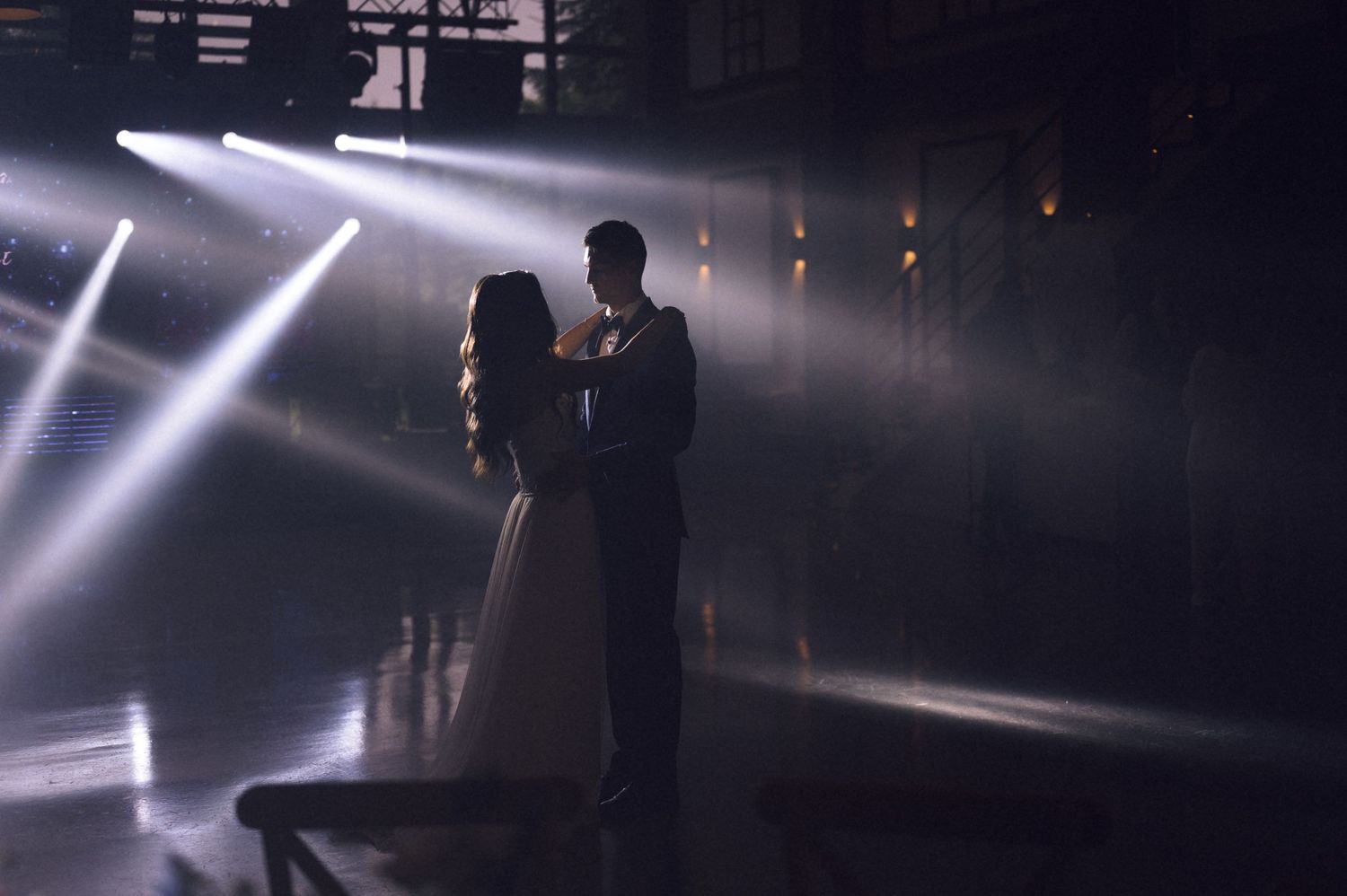 Couple sharing romantic first dance in dramatic spotlight beams on dark industrial venue dance floor.