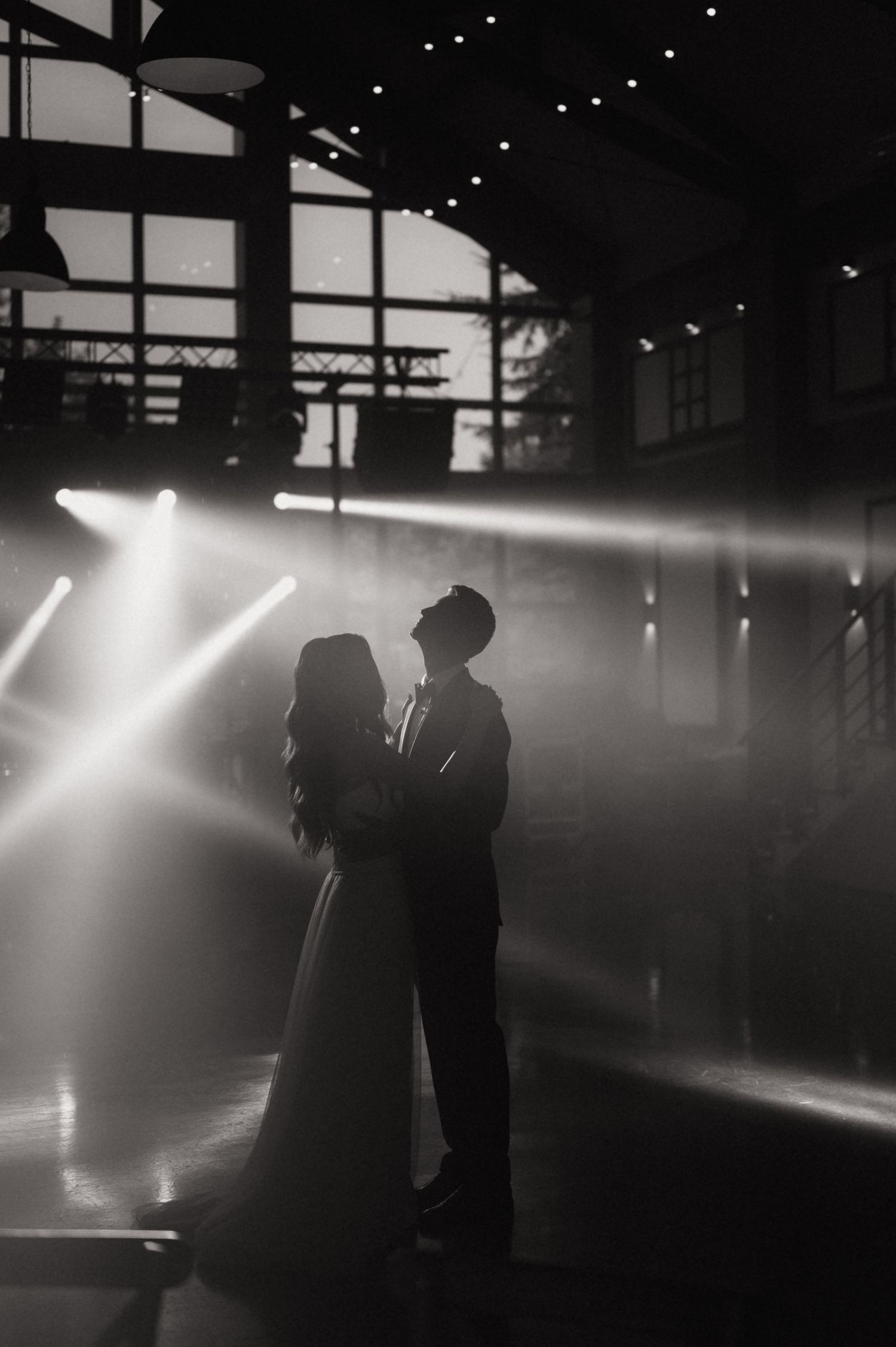 Black and white silhouette of a bride and groom sharing a romantic moment under dramatic light beams at a wedding venue.
