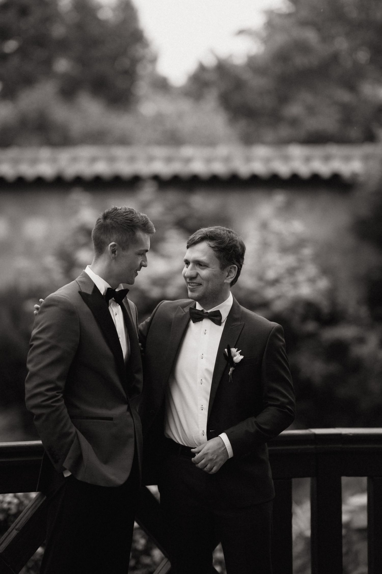 Two grooms in black tuxedos with bow ties share a tender moment on a wooden bridge in a black and white wedding photo.