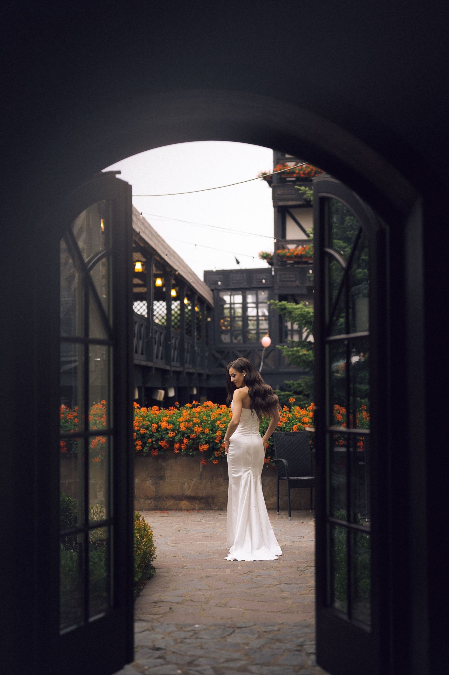 Bride in white gown standing in courtyard framed by arched French doors with orange flowers.