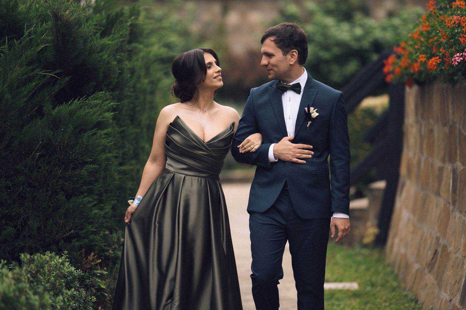 Elegant couple in formal attire walking together outdoors, man in tuxedo and woman in olive green gown.