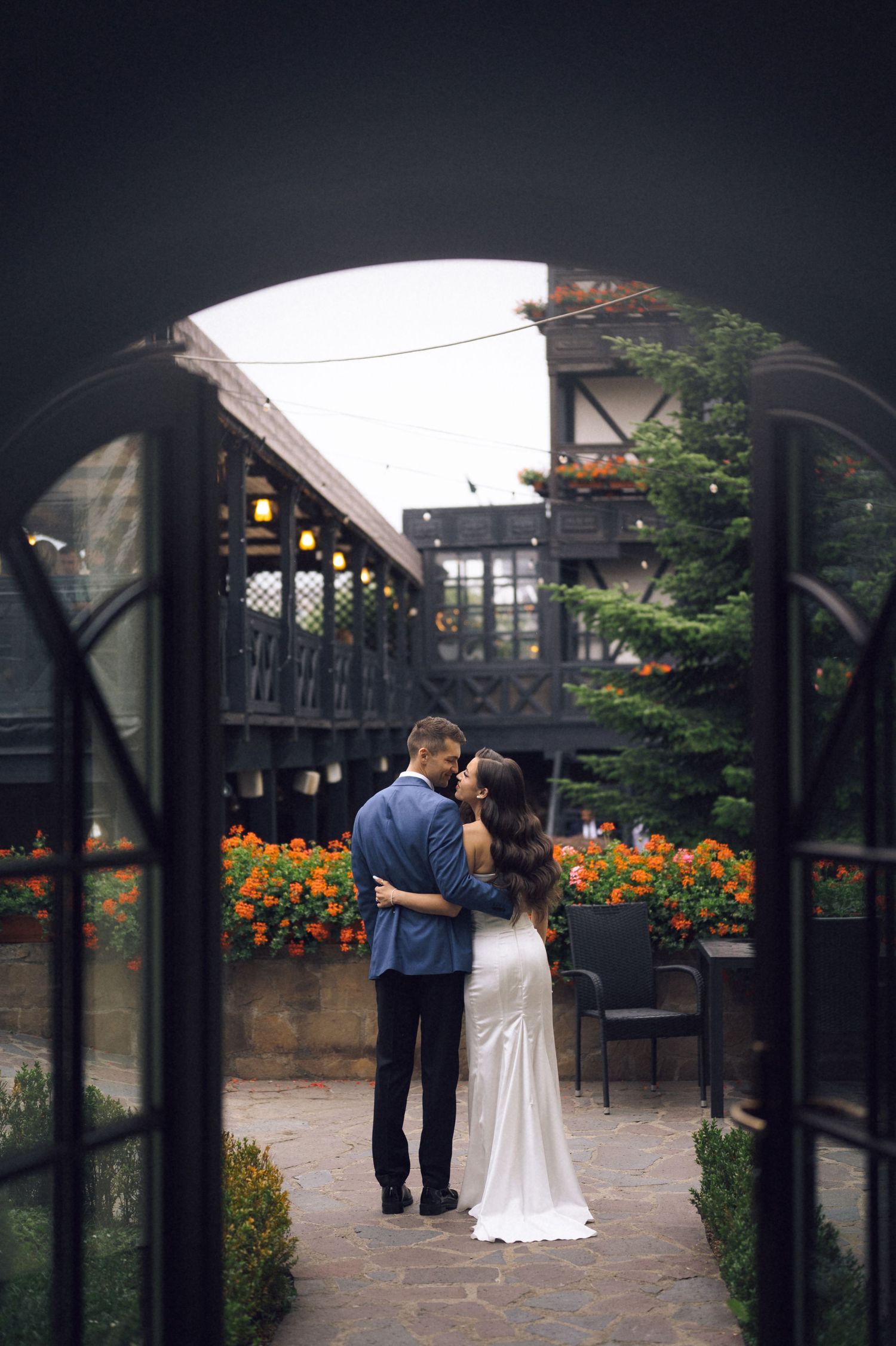 Couple embracing in flower-filled courtyard viewed through elegant arched French doors.