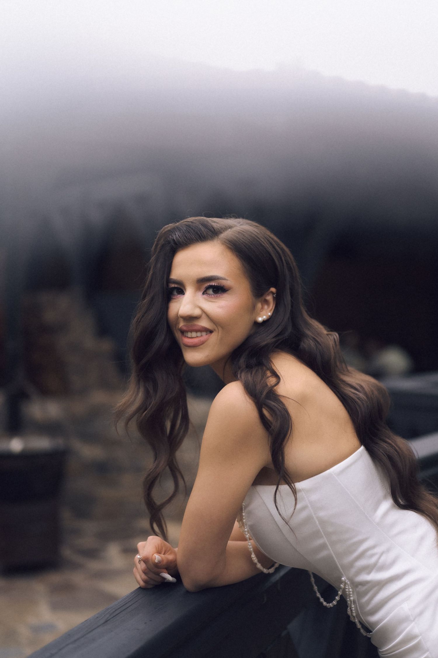 Young woman in white dress with long wavy hair smiles over her shoulder while leaning on a railing in misty scenery.