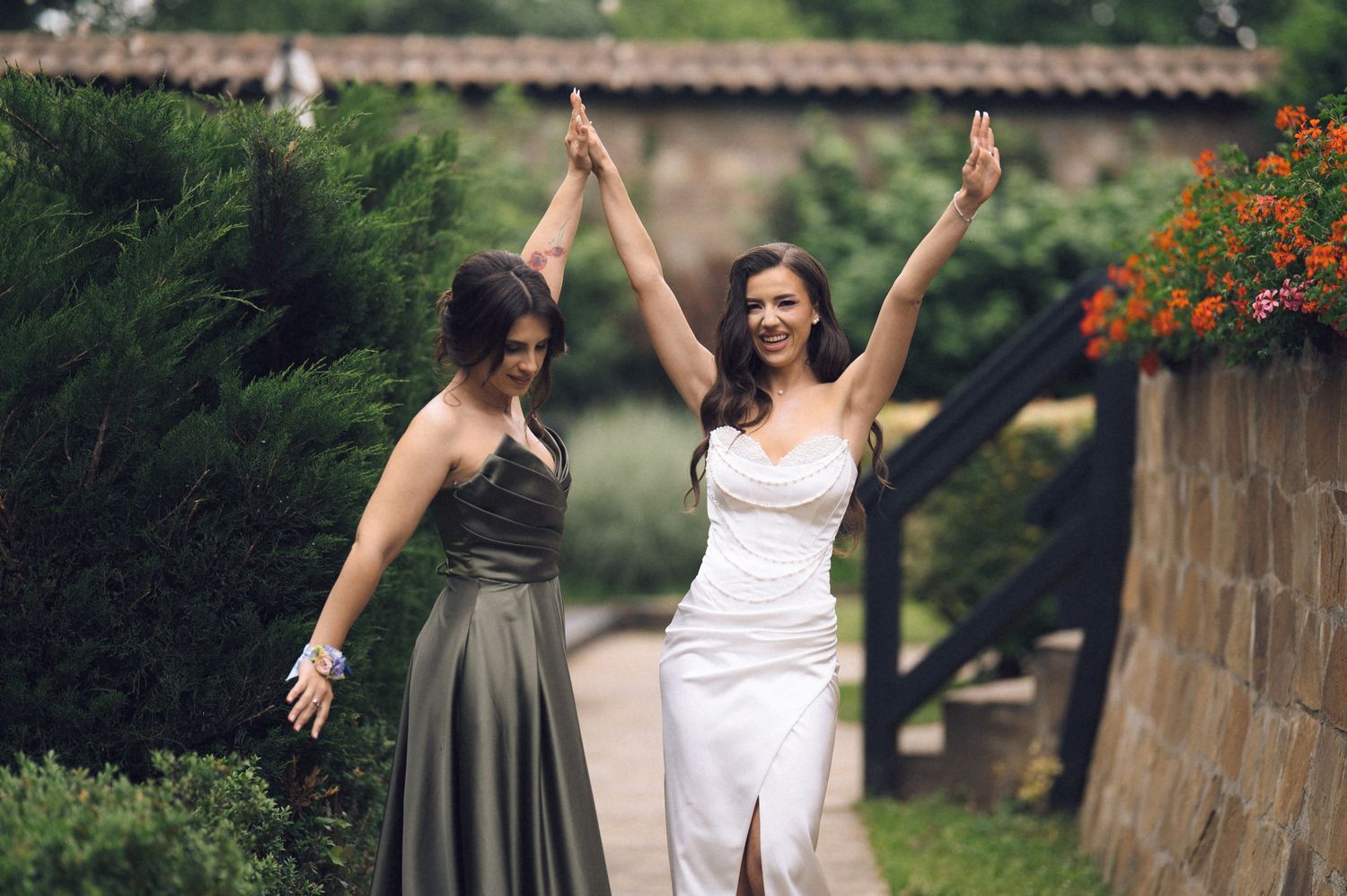 Bride in white gown and bridesmaid in gray dress celebrate with raised arms on a garden path.