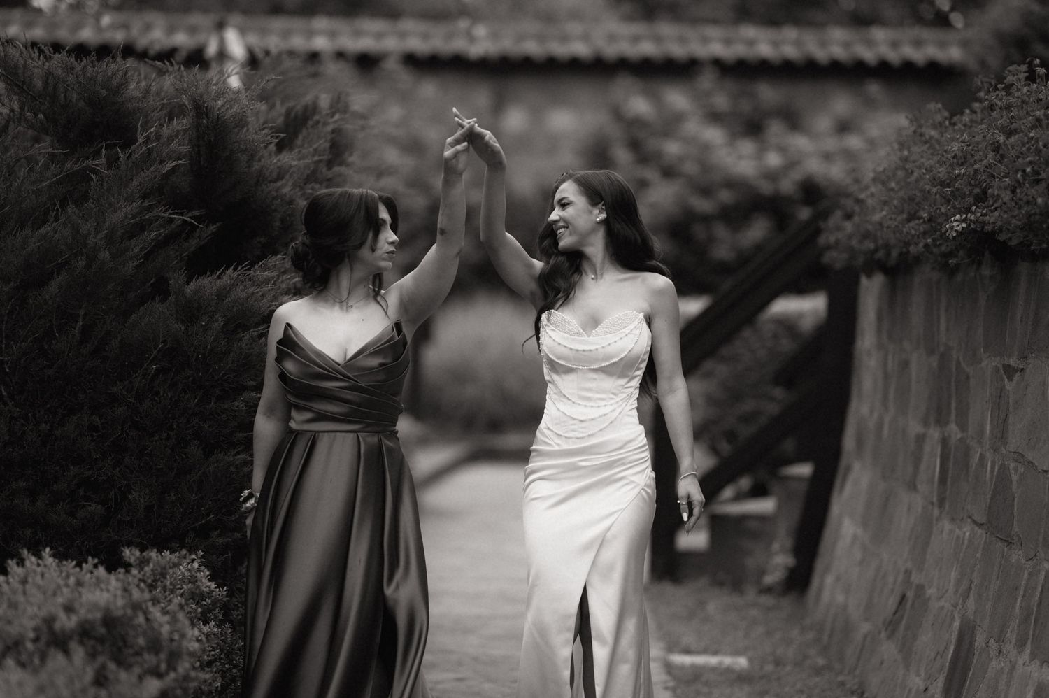 Two women in elegant gowns walk together, one in white bridal dress, one in satin, holding hands raised in celebration.